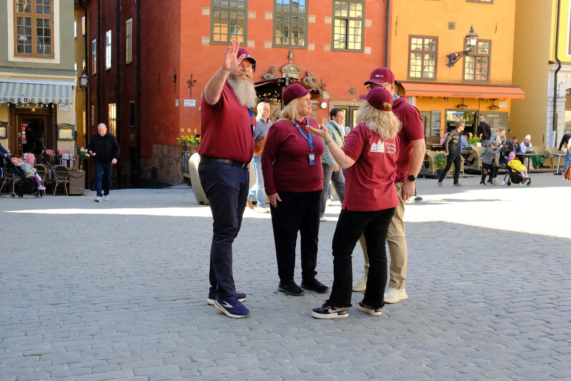 En grupp i röda tröjor, en med texten "STOCKHOLM CITY GUIDE", på kullerstensgata i stadsmiljö. En man vinkar